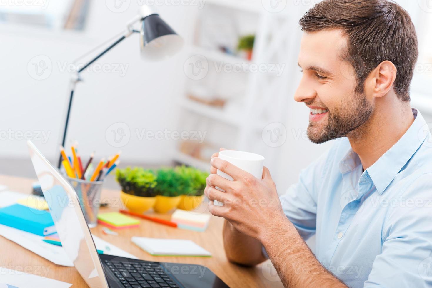 trabajando con placer. apuesto joven en camisa sosteniendo una taza de café y sonriendo mientras está sentado en su lugar de trabajo foto