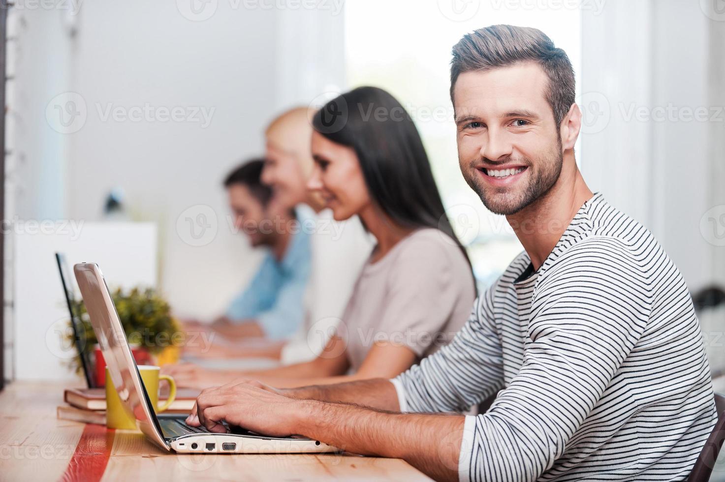 I love my job Group of cheerful business people in smart casual wear working at their laptops while handsome man looking at camera and smiling photo