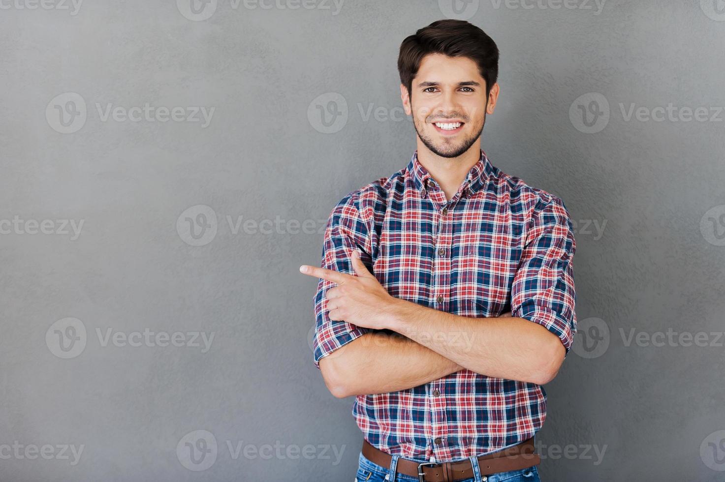 Pointing copy space. Handsome young man pointing away and smiling while standing against grey background photo