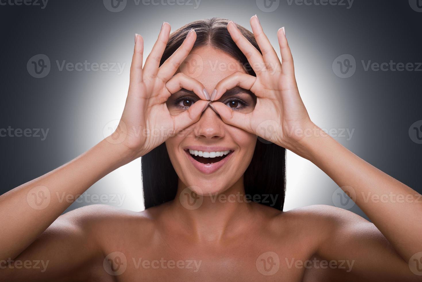 Time for fun. Portrait of cheerful young shirtless woman looking at camera through fingers while standing against grey background photo