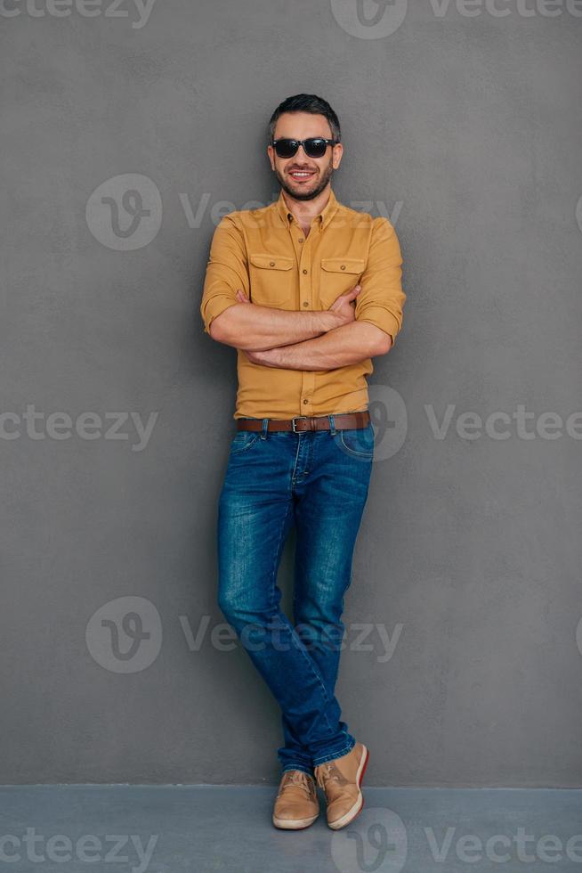 casualmente guapo. longitud completa de un hombre maduro confiado en gafas de sol con los brazos cruzados y mirando a la cámara con una sonrisa mientras se enfrenta a un fondo gris foto
