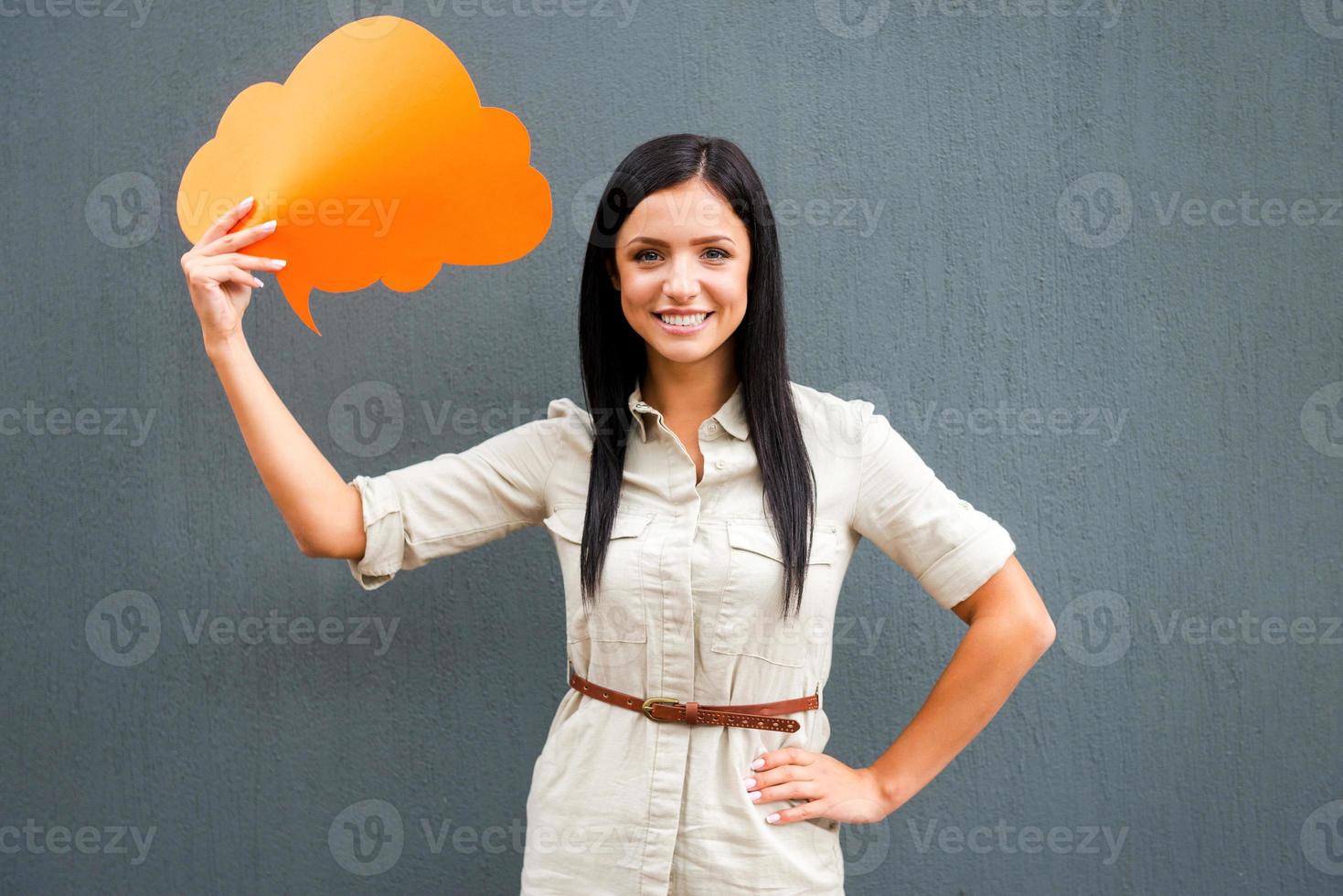 Put your thoughts in that cloud. Smiling young woman holding empty speech bubble and looking at camera while standing against grey background photo