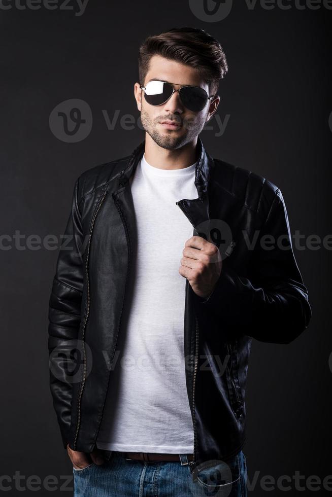 Trendy handsome. Handsome young man adjusting his leather jacket and looking at camera while standing against black background photo