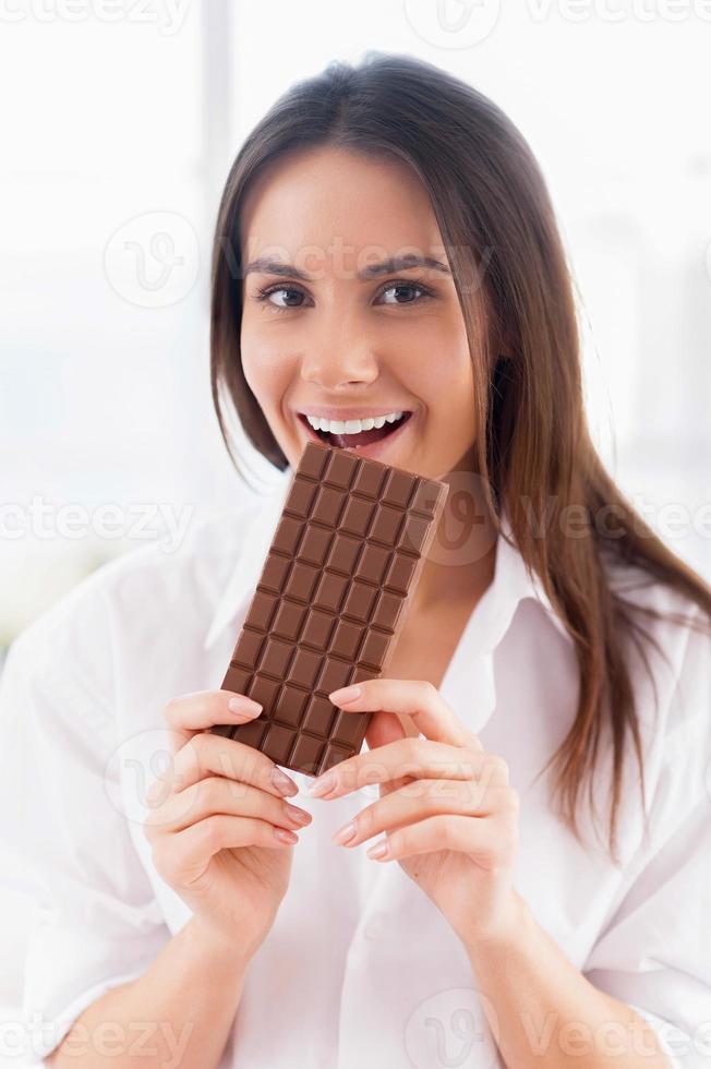 Girl enjoying chocolate. Cheerful young smiling woman in white shirt eating chocolate bar and looking at camera photo