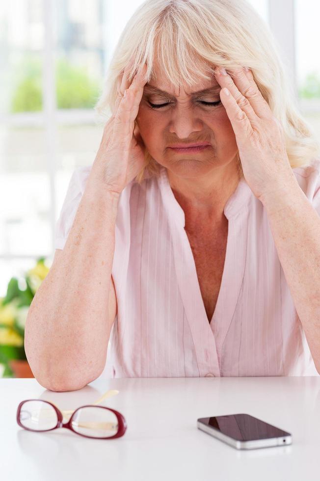 She needs help. Depressed senior woman holding head in hands and keeping eyes closed while sitting at the table with mobile phone laying on it photo