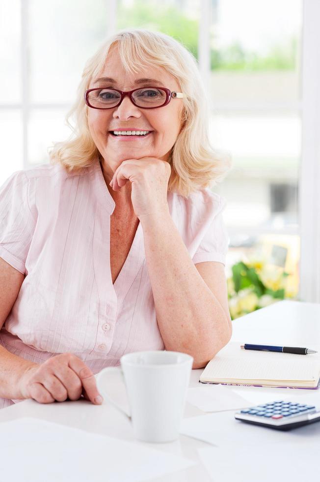 Getting finances straight. Happy senior woman holding hand on chin and smiling at camera while sitting at the table photo