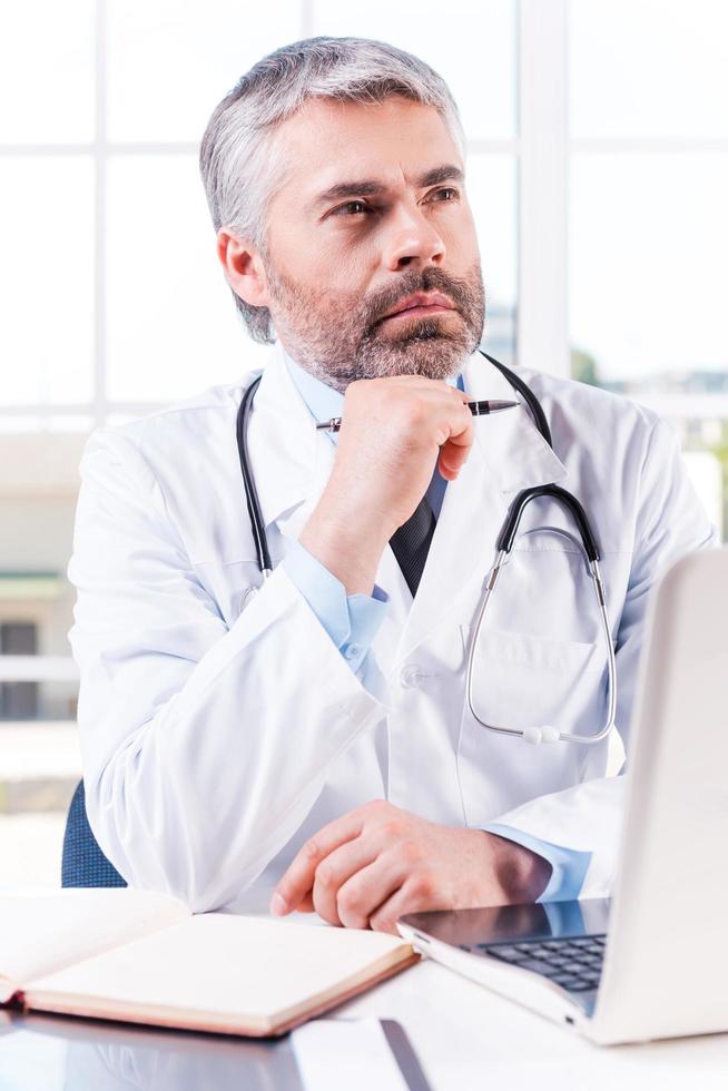 Thoughtful doctor. Thoughtful mature grey hair doctor holding hand on chin and looking away while sitting at his working place photo