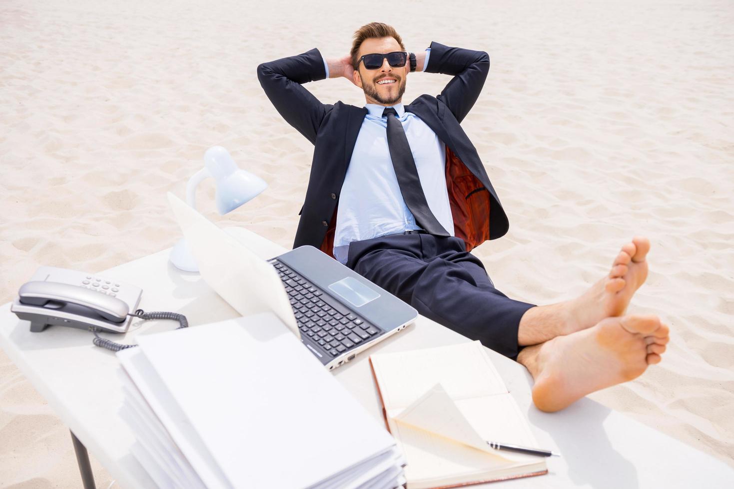 Total relaxation. Top view of relaxed young man in formalwear and sunglasses holding hands behind head and holding his feet on the table standing on sand photo