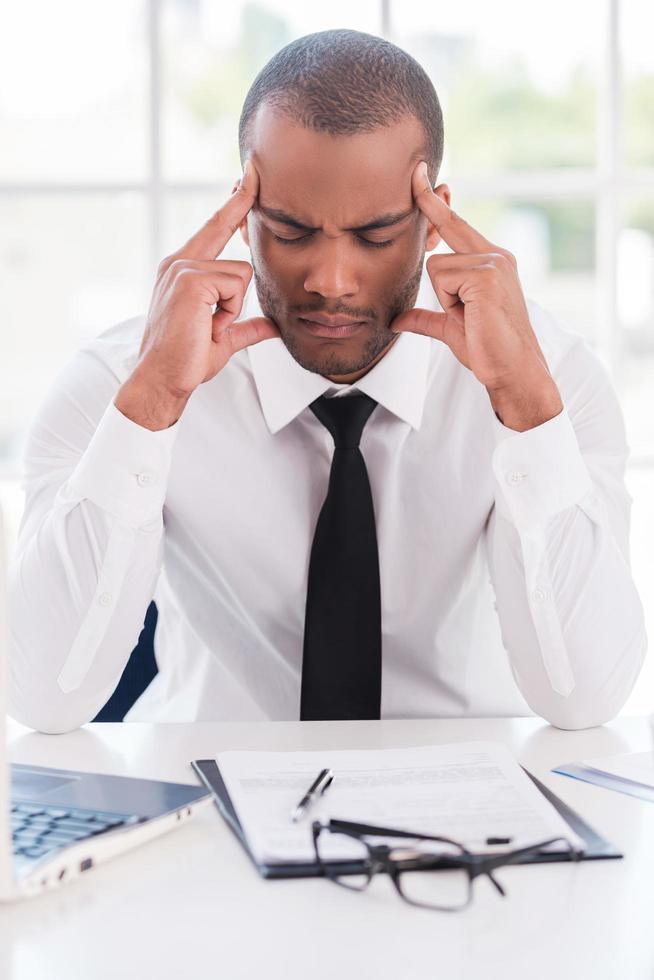 estresado y cansado. joven africano deprimido con ropa formal sosteniendo la cabeza en las manos y manteniendo los ojos cerrados mientras se sienta en su lugar de trabajo foto