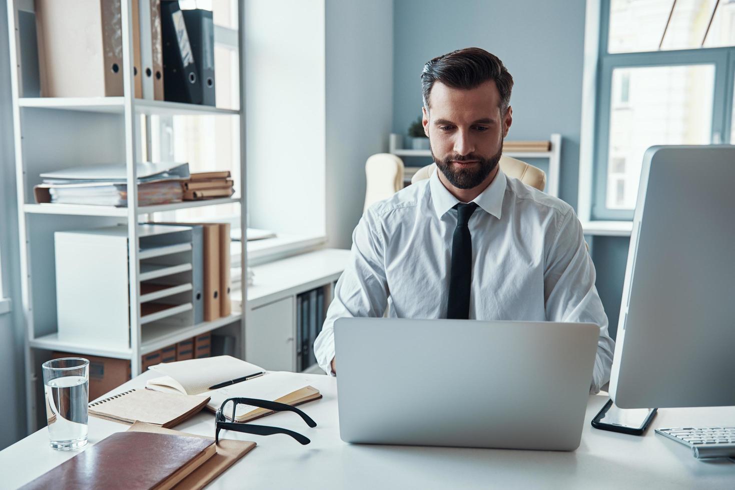 joven concentrado en camisa y corbata trabajando usando computadora mientras está sentado en la oficina foto