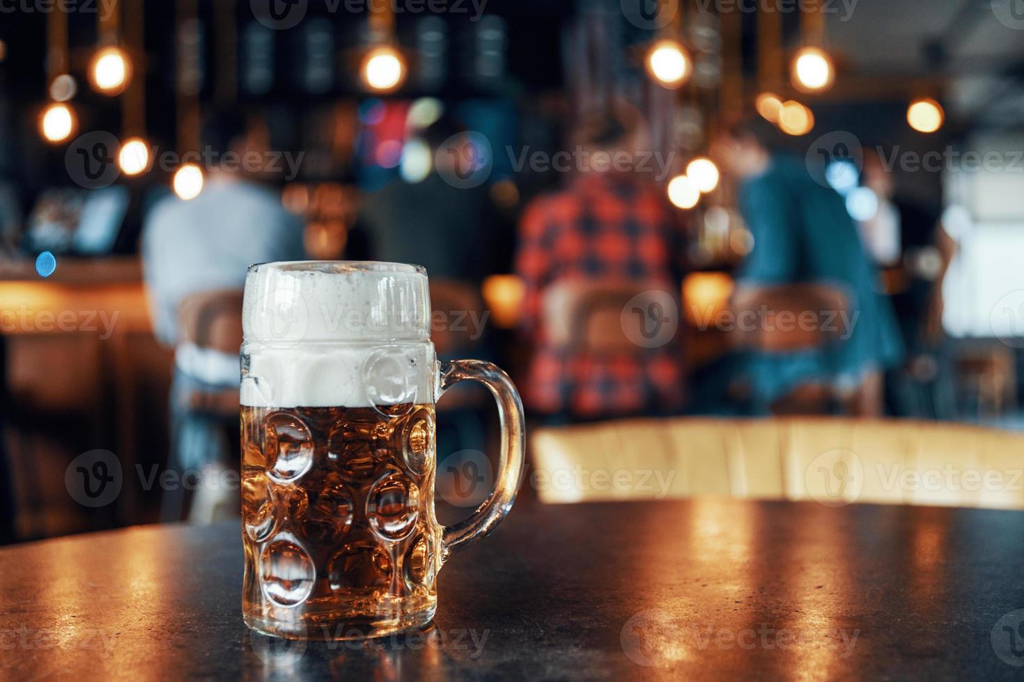 Pint glass of cold fresh beer with foam standing on the table in pub with people on the background photo
