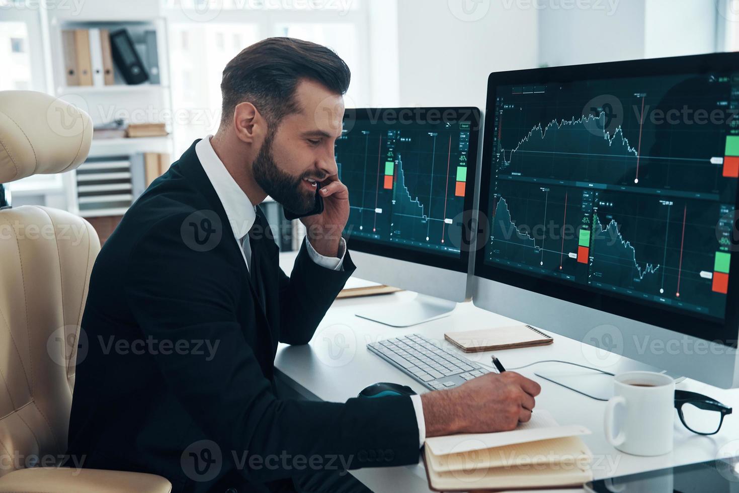 Concentrated young man in shirt and tie writing something down and talking on the phone while working in the office photo