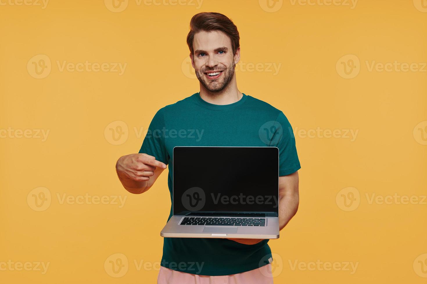 Handsome young smiling man in casual clothing carrying laptop and pointing monitor photo