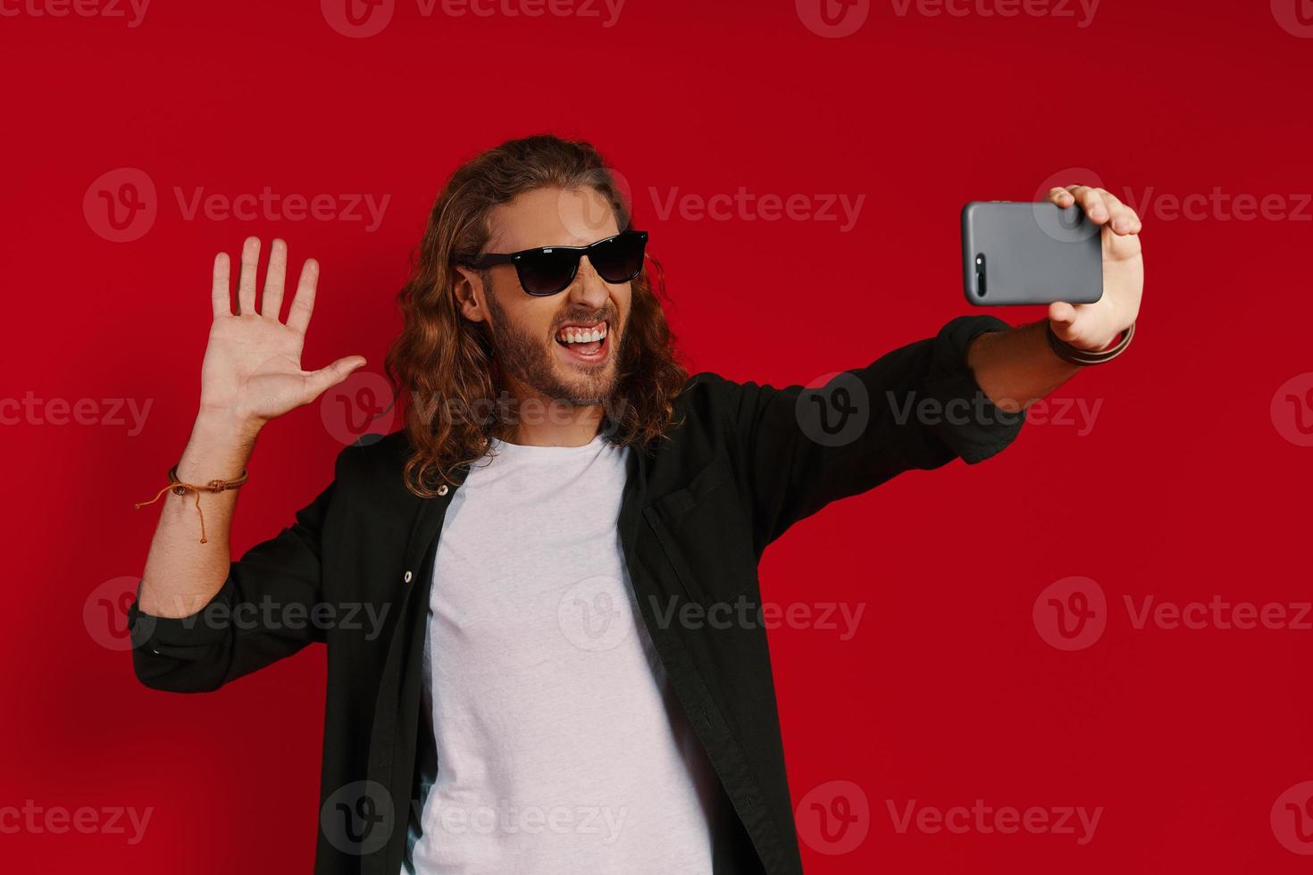 Cheerful young man in casual clothing taking selfie and smiling while standing against red background photo