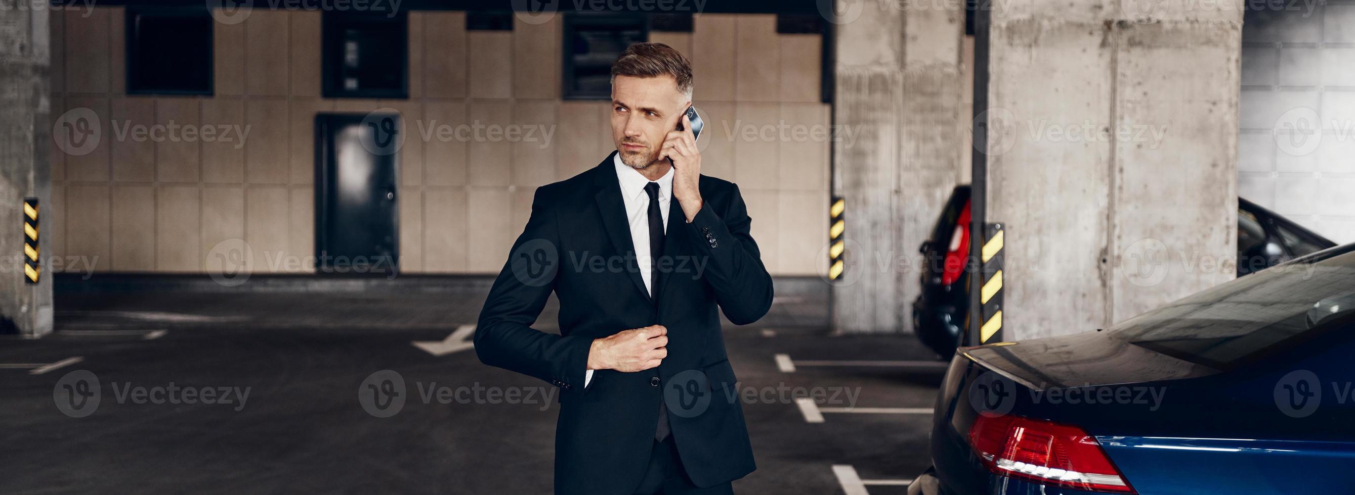 Confident mature businessman talking on mobile phone while walking by parking lot photo