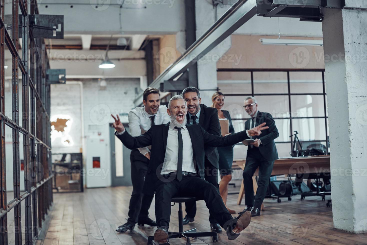 Playful businessmen pushing their boss on the office chair while running in the hallway photo