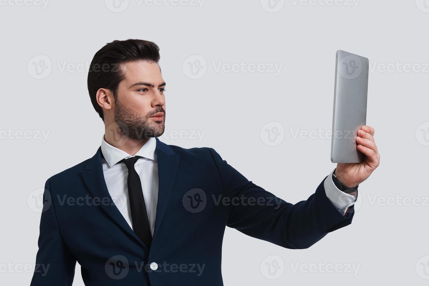 Hi Good looking young man in full suit holding digital tablet and looking at it while standing against grey background photo