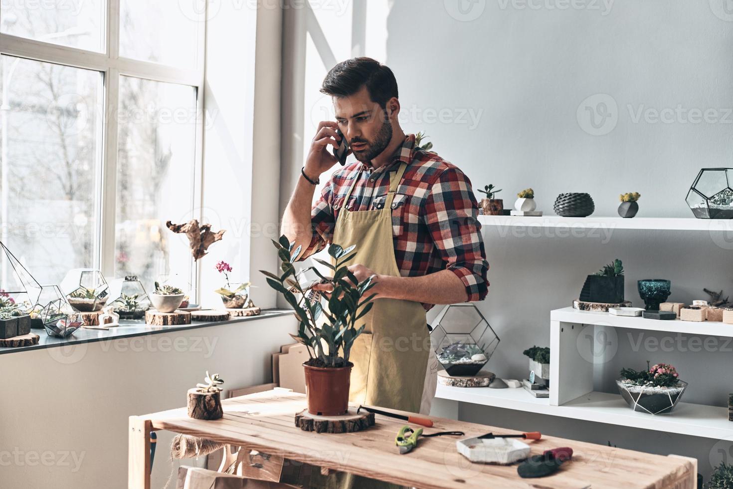 Taking order. Handsome young man in apron talking on the phone while standing in small garden center photo