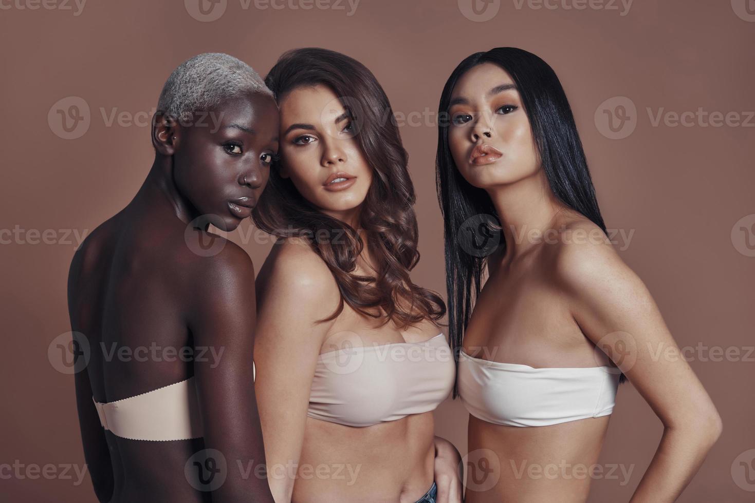 Beautiful females. Three attractive young women looking at camera while standing against brown background photo