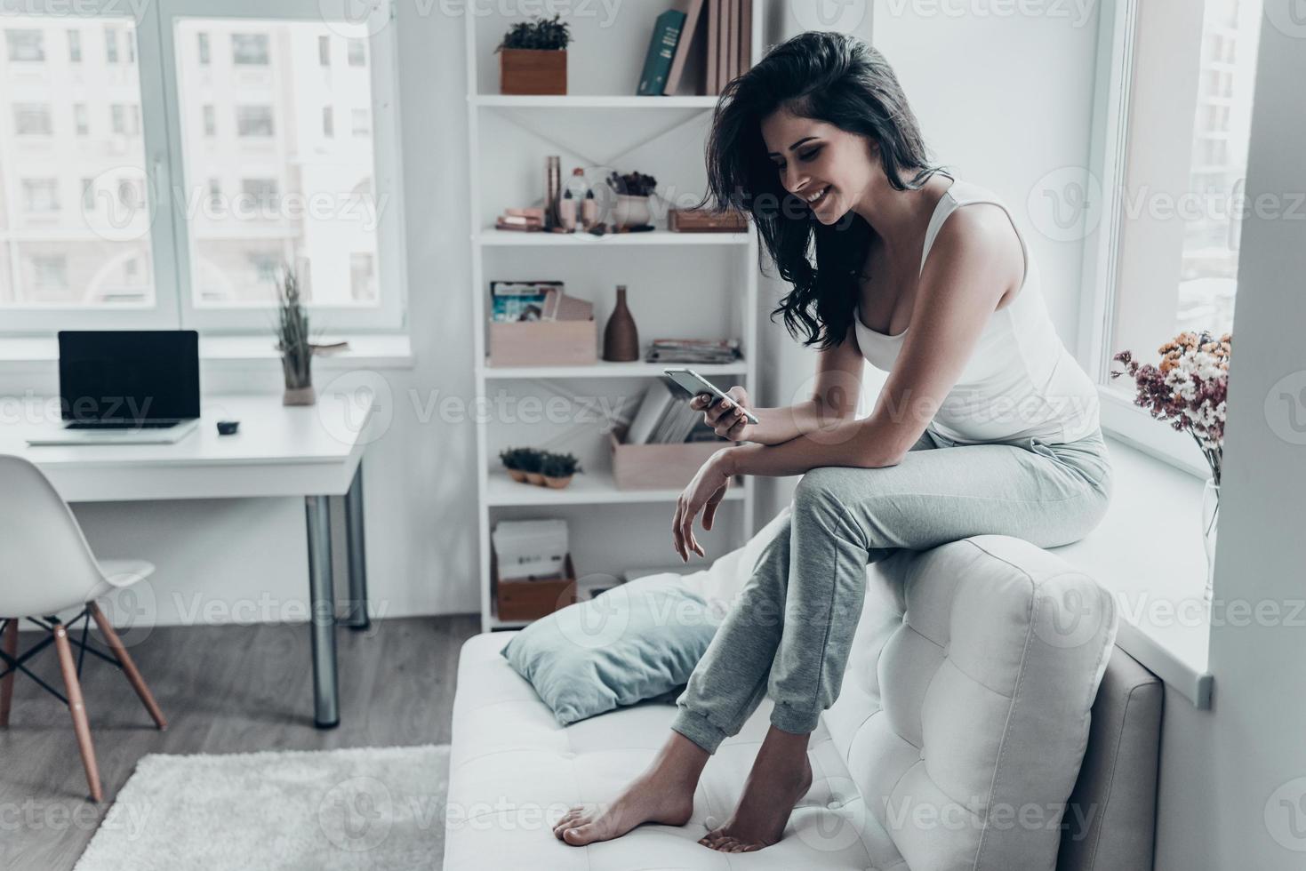 Receiving lovely messages. Attractive young woman using her smart phone and smiling while sitting on window sill photo