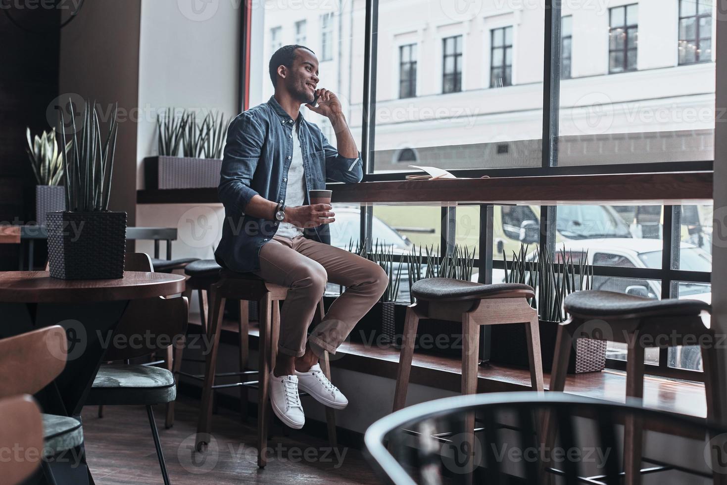 apuesto joven hablando en su teléfono inteligente con una sonrisa y sosteniendo una taza desechable mientras está sentado en la cafetería foto