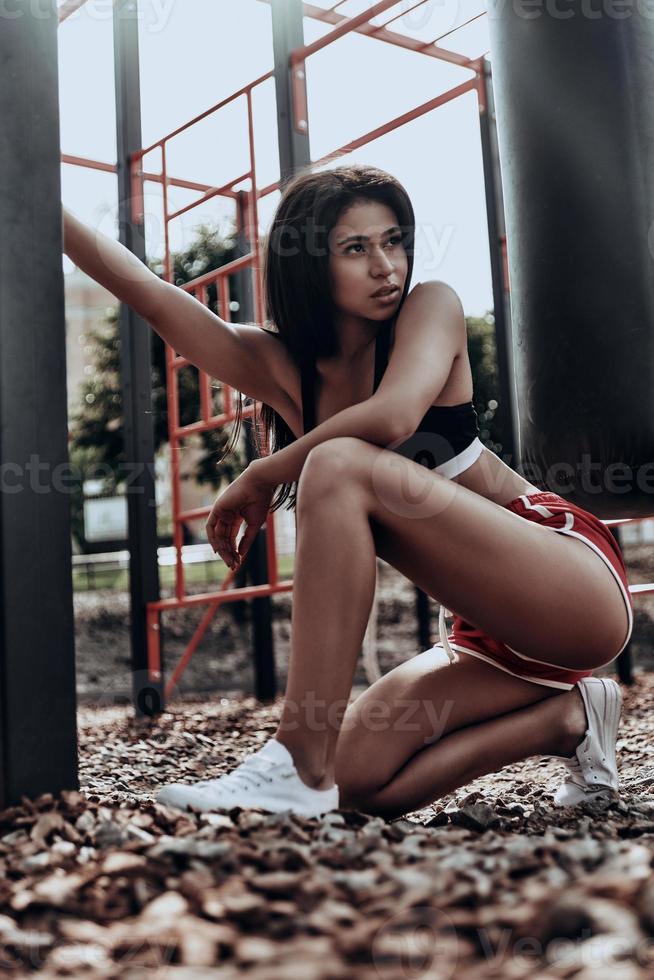 belleza deportiva. atractiva mujer joven con ropa deportiva mirando hacia otro lado mientras se agacha en el parque de gimnasia al aire libre foto