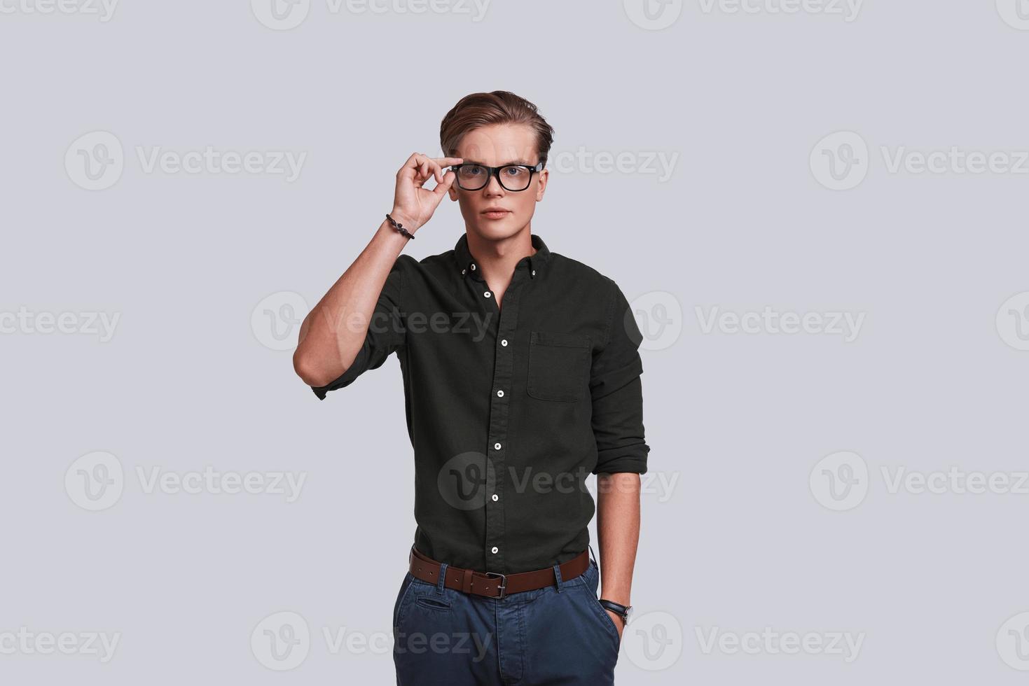 Taking everything seriously. Good looking young man adjusting his eyewear and keeping hand in pocket while standing against grey background photo