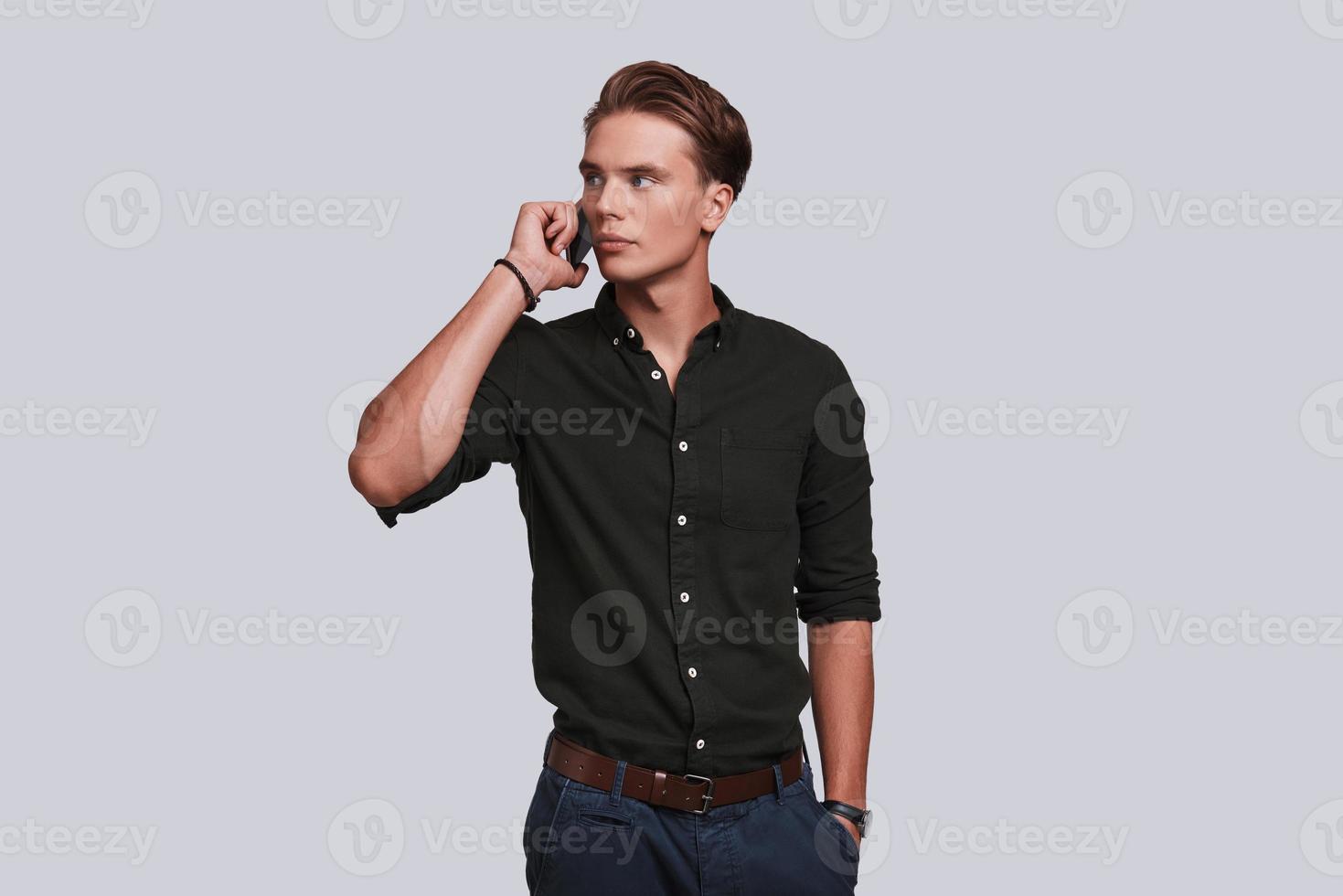 Serious young man talking on smart phone and keeping hand in pocket while standing against grey background photo