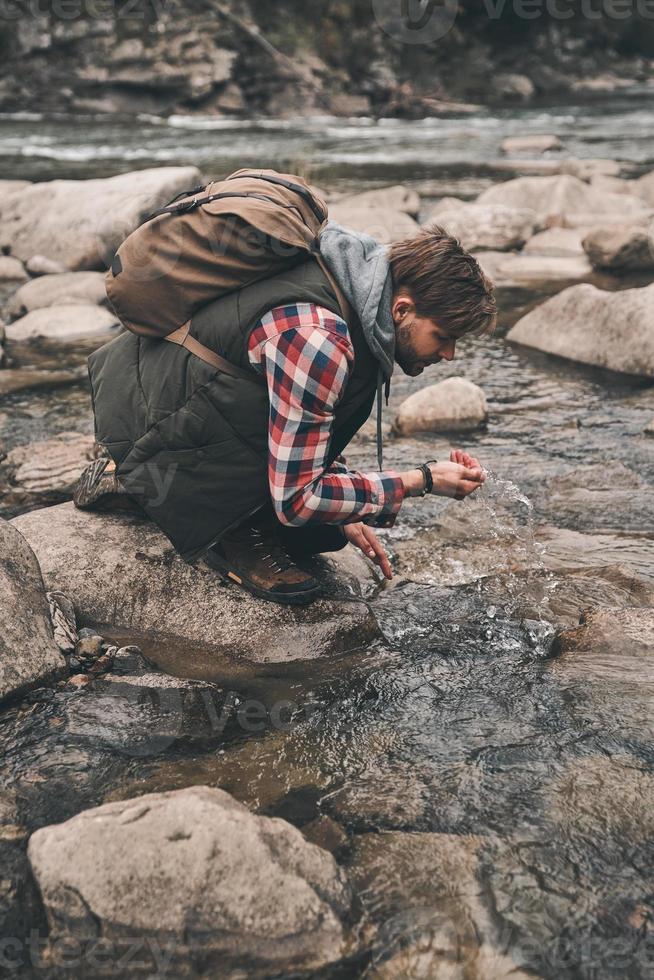 Refreshing with cold water. Handsome young modern man drinking water from the river while hiking in mountains photo