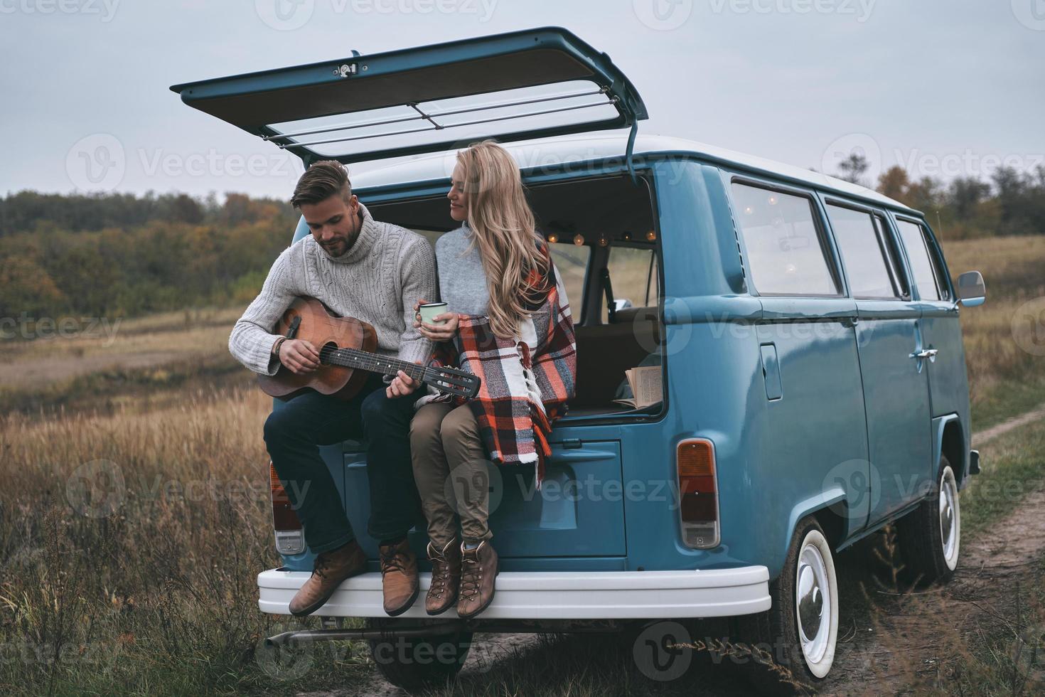 Romantic couple.  Handsome young man playing guitar for his beautiful girlfriend while sitting in the trunk of blue retro style mini van photo