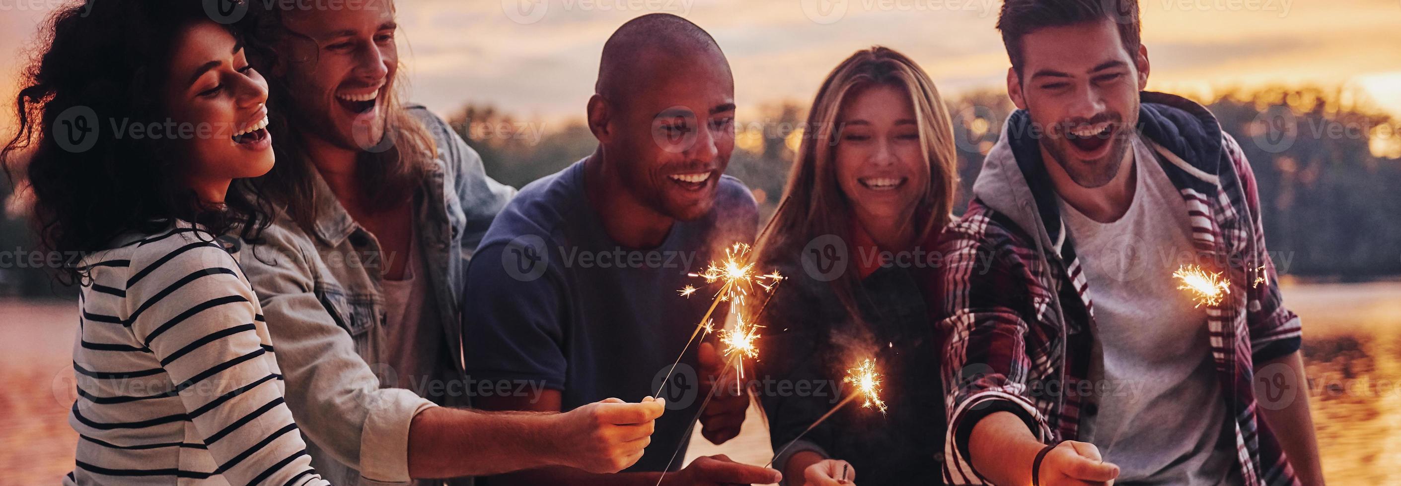 divirtiéndose. grupo de jóvenes con ropa informal sonriendo y sosteniendo chispas mientras están de pie en el muelle foto