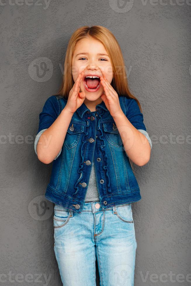Can you hear me now Cheerful little girl keeping mouth open and looking at camera while standing against grey background photo