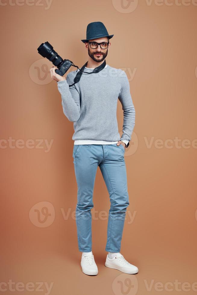 Full length of charming young man in casual wear smiling and looking at camera while standing against brown background photo