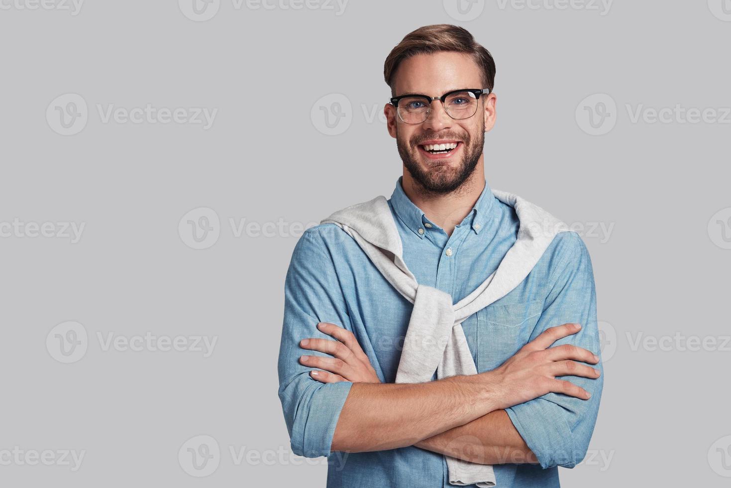 Confident in his style.  Good looking young man keeping arms crossed and looking at camera with smile while standing against grey background photo