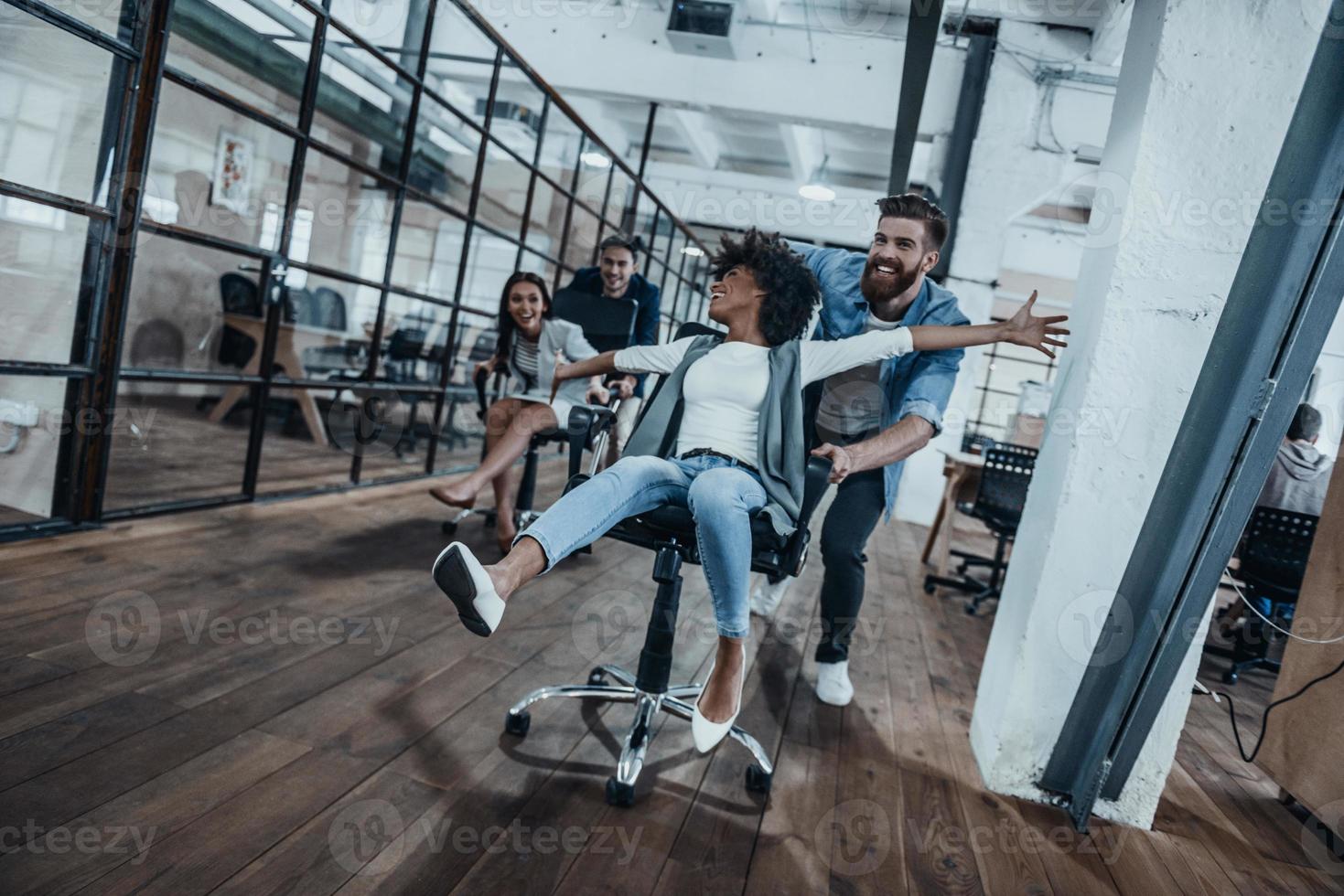 Four young cheerful business people in smart casual wear having fun while racing on office chairs and smiling photo