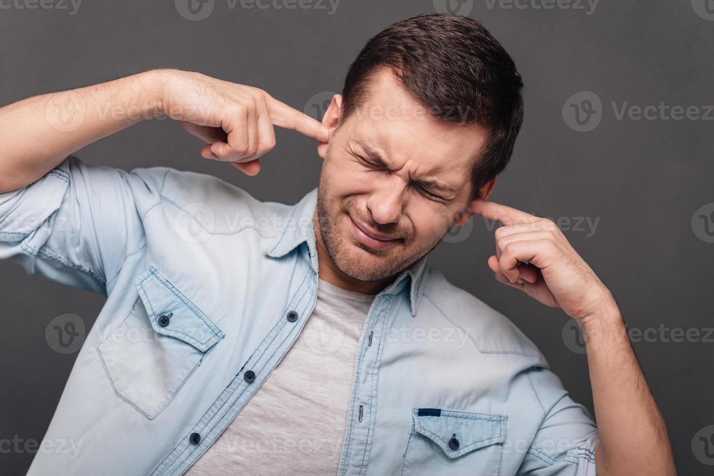 Too loud Young handsome man covering ears by hands and keeping eyes closed while standing against grey background photo