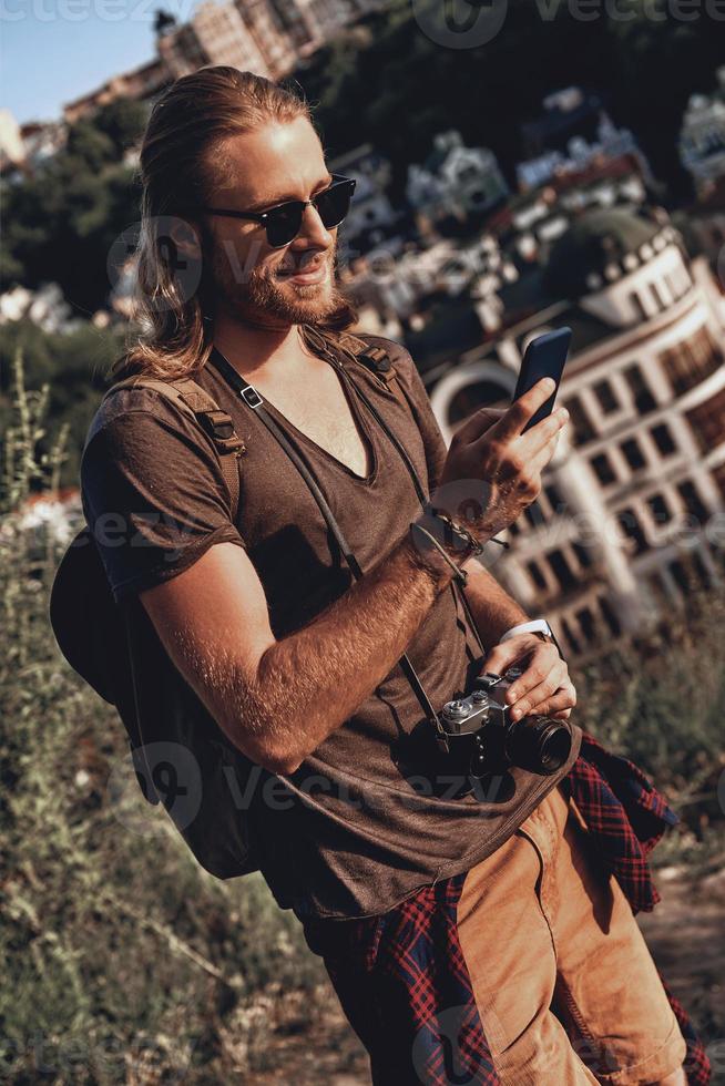en línea en el mensajero. joven con ropa informal usando un teléfono inteligente y sonriendo mientras está de pie en la colina al aire libre foto