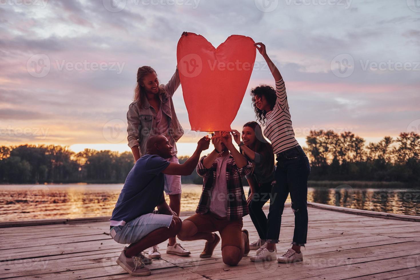 Heart shaped lantern. Full length of young people in casual wear lighting up sky lantern while standing on the pier photo