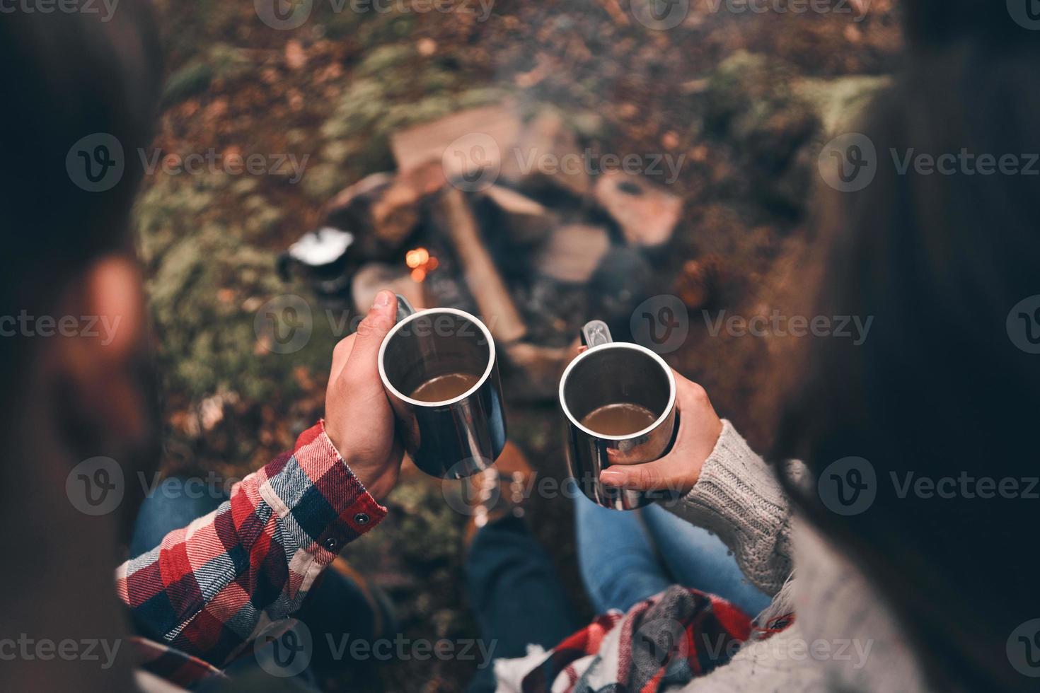 tomarse un tiempo para descansar. cerrar la vista superior de una pareja joven sosteniendo tazas mientras se calienta cerca de la fogata foto