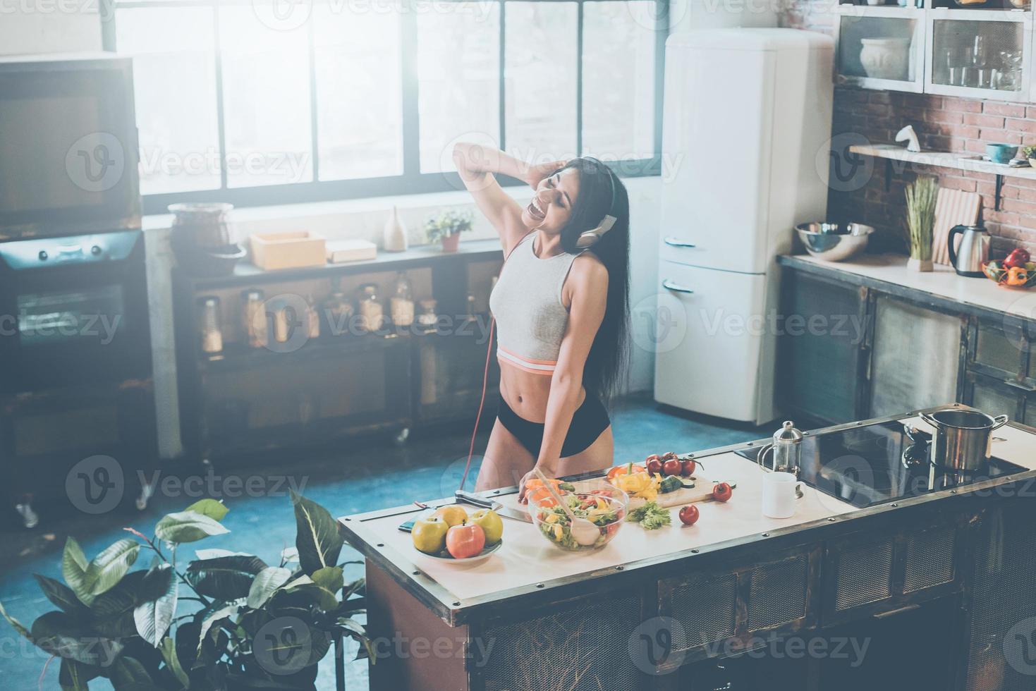 Cooking with nice music. Top view of beautiful young African woman in headphones dancing and expressing positivity while preparing food in kitchen at home photo