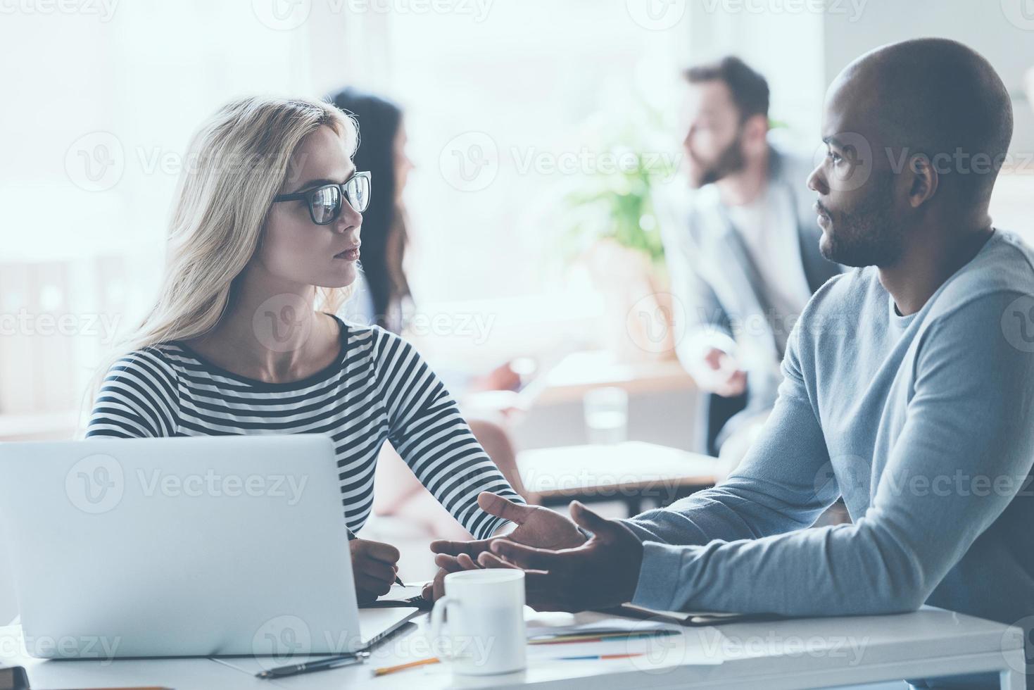 Discussing some business issues. Two young business people discussing something while sitting at the office desk together while their colleagues sitting in the background photo