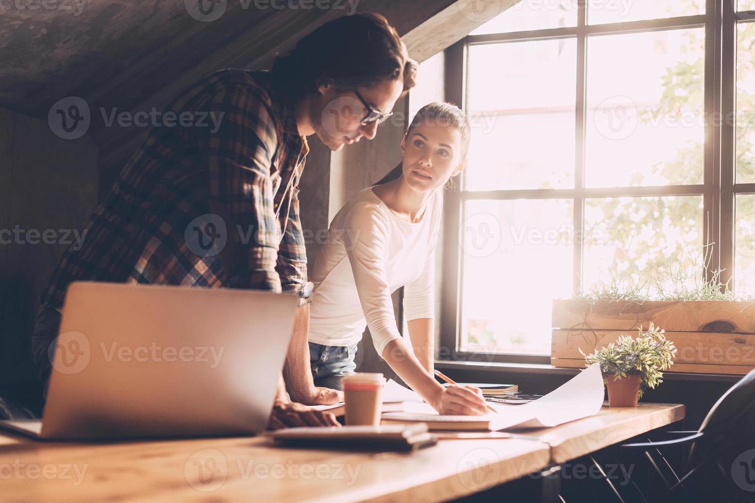 Finding solution together. Confident young man and woman in casual wear pointing blueprint and communicating while both standing near wooden desk in creative office photo