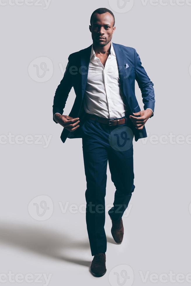 Confidence in every move. Full length of handsome young African man in full suit adjsuting his jacket while walking ahead and being in front of grey background photo