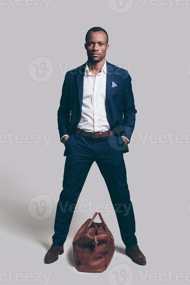 Me and my bag. Full length of handsome young African man in full suit holding hands in pockets and looking at camera while standing against grey background with brown leather bag laying near him photo