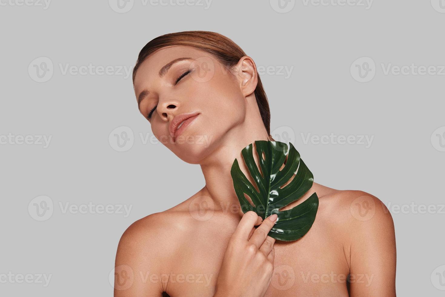 Time for yourself. Attractive young woman keeping eyes closed and covering with leaf while standing against grey background photo