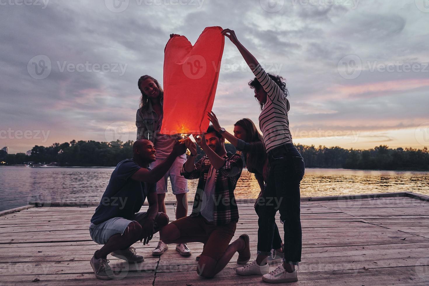 Heart shaped lantern. Full length of young people in casual wear lighting up sky lantern while standing on the pier photo