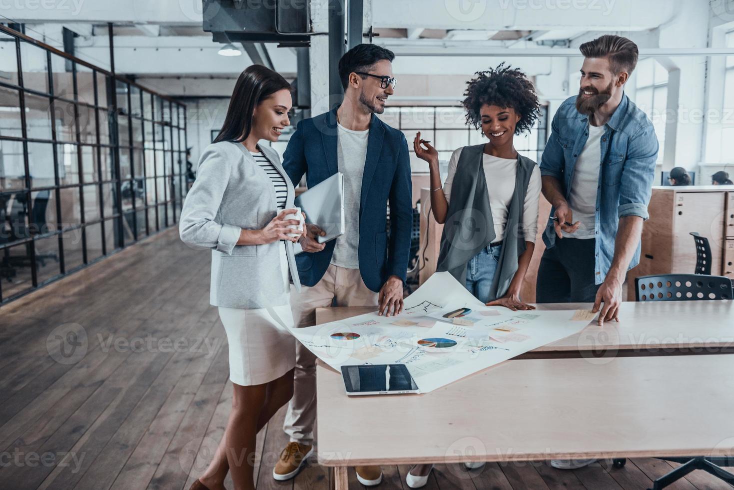 Best team ever. Group of young business people in smart casual wear working together and smiling while standing near the wooden desk in office photo