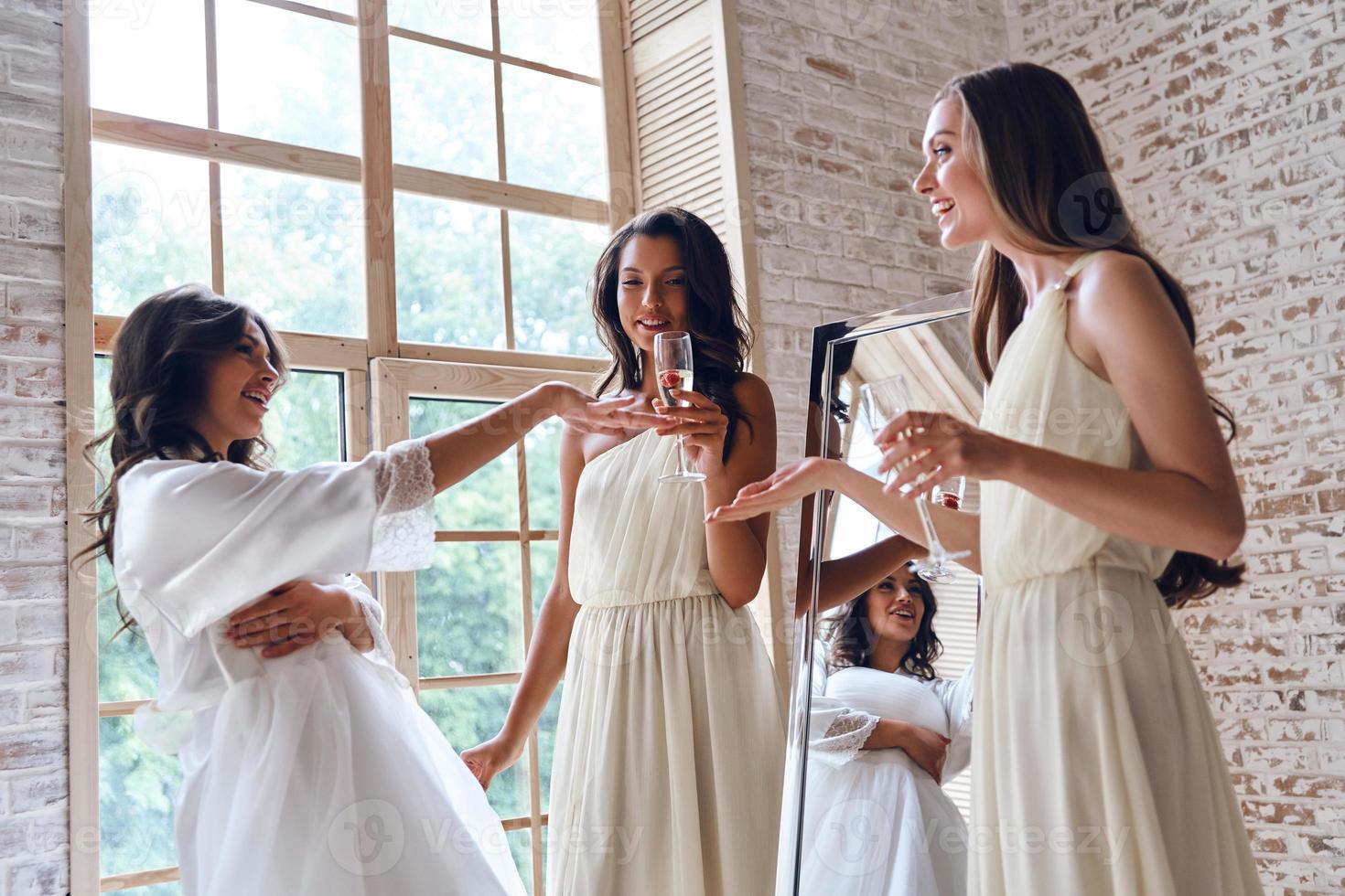 Two attractive young women smiling while looking at brides wedding ring while standing near the window together photo