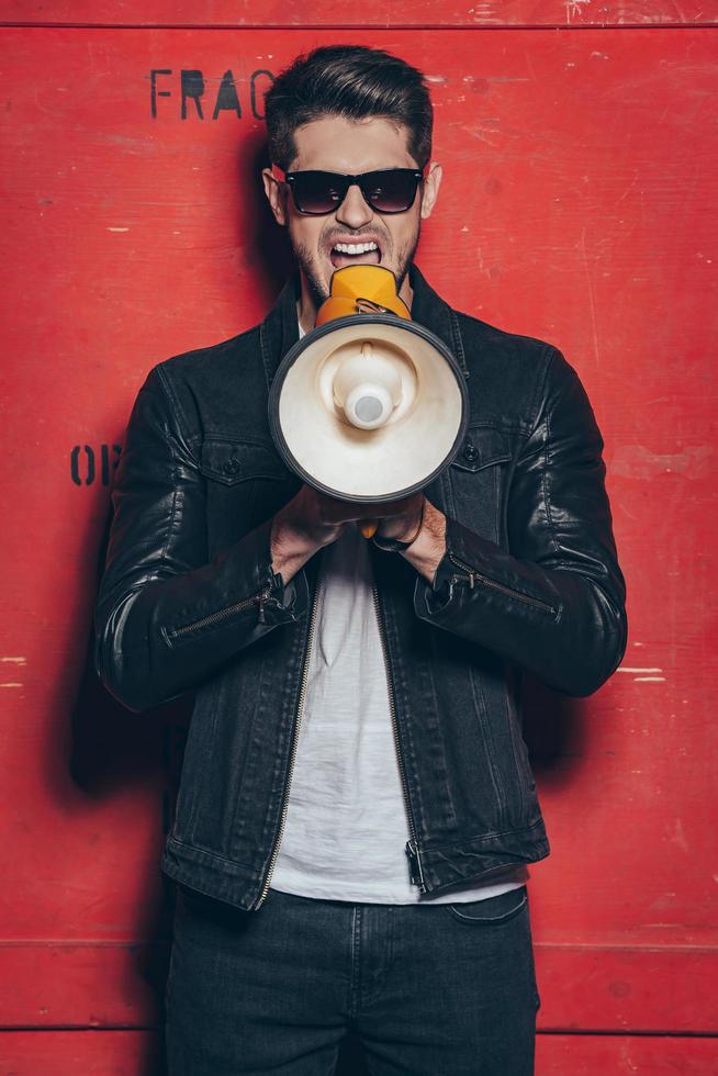 Stylish announcement. Handsome young man in sunglasses holding megaphone and keeping mouth open while standing against red background photo
