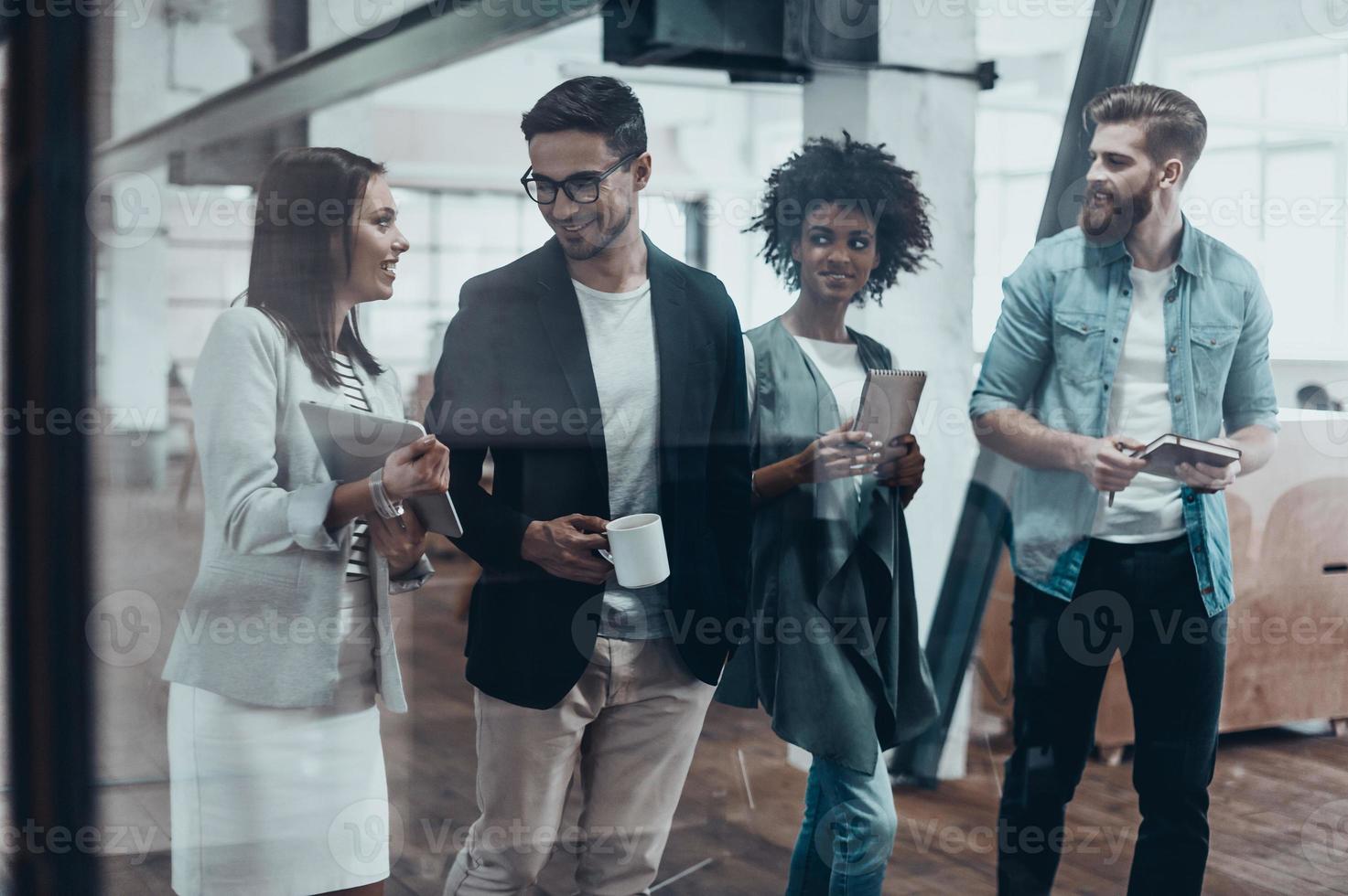 Working beyond the boarders of a desk. Group of young people in smart casual wear talking and smiling while walking together behind the glass wall in the creative office photo